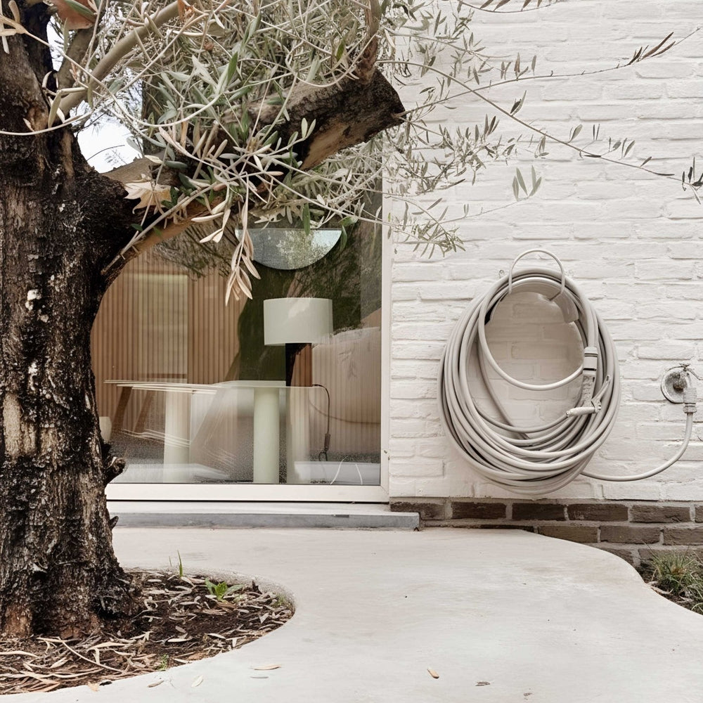 Design Gartenschlauch Gentle Greige von LIVLIG, aufgerollt an einer weißen Backsteinwand mit hellgrauer Schlauchhalterung.