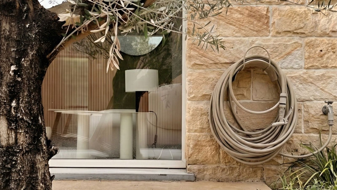 Nahaufnahme einer Außenwand aus Sandstein mit einem aufgerollten Gartenschlauch, daneben ein Baum und ein Fenster mit Tisch und Lampe.