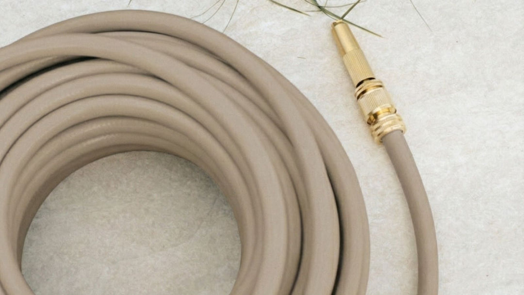 Auf einer beigen Oberfläche liegt ein zusammengerollter, beiger Gartenschlauch mit goldfarbenem Anschlussstück rechts. Wenige grüne Blätter liegen darüber.