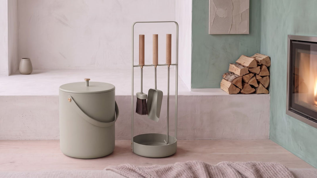 Sage green fireplace tools set with matching bucket, wooden handles, and metal stand, beside stacked firewood and lit fireplace.