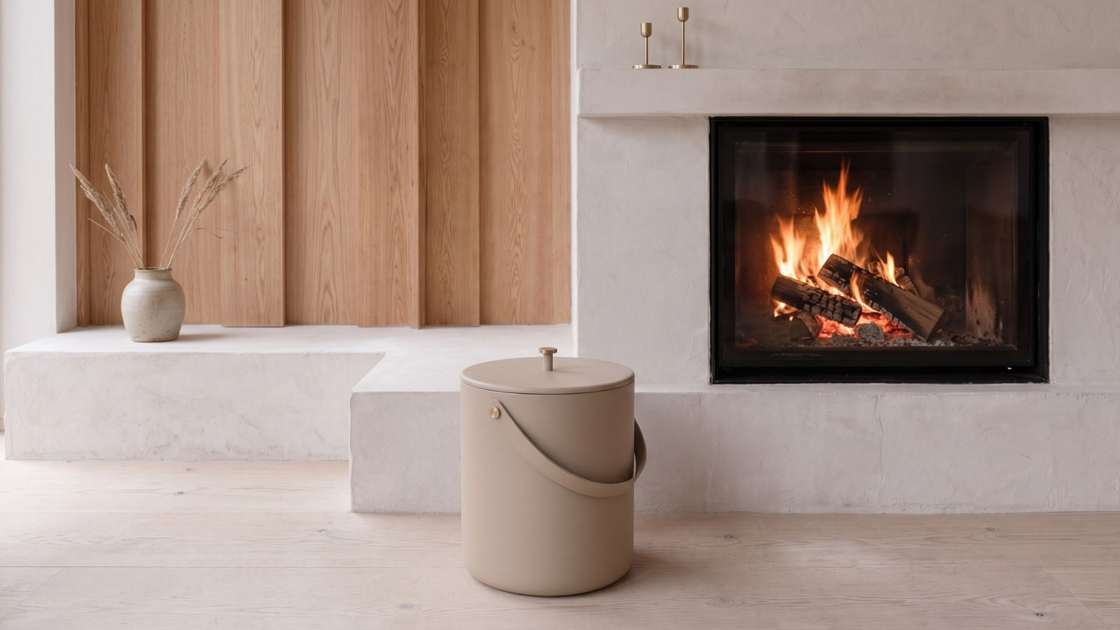 Modern living room with lit fireplace, beige cylindrical bucket, wooden paneling, and dried grass in vase on concrete ledge.