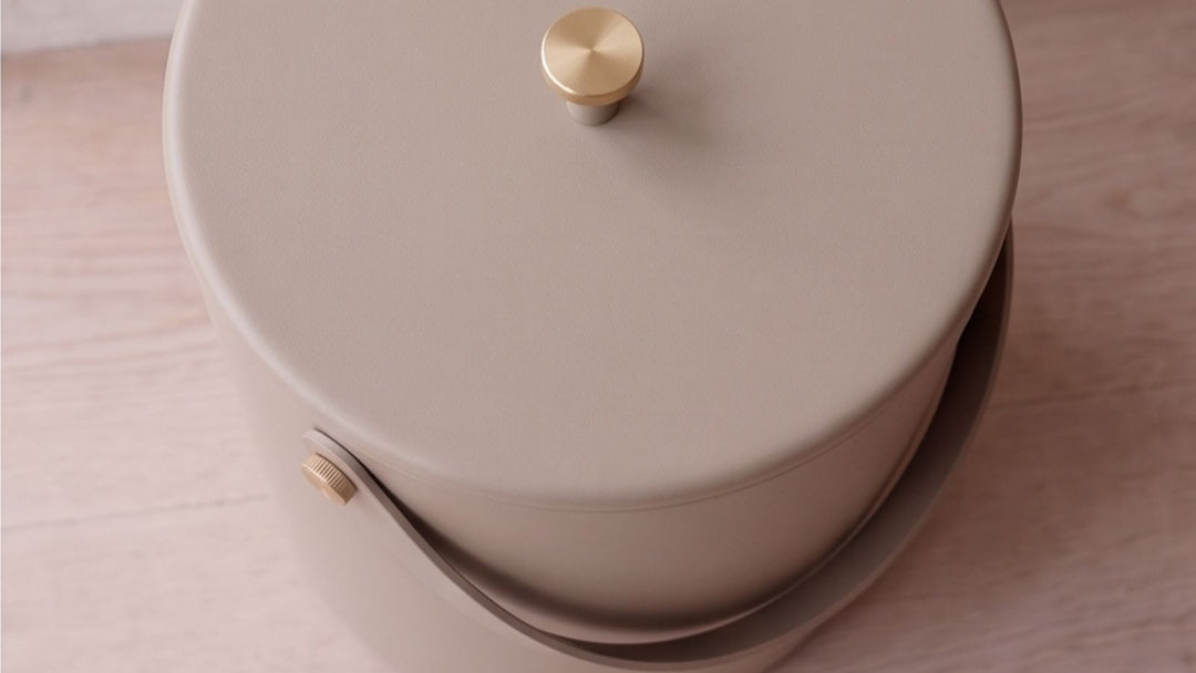 Beige cylindrical container with gold knob on lid and handle, resting on light wood surface.