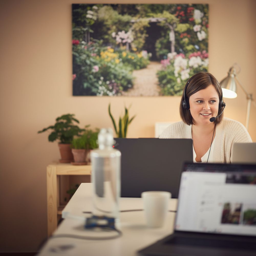Eine lächelnde Frau mit Headset arbeitet an einem Laptop. Im Hintergrund ein Blumenbild und Pflanzen auf einem Regal.