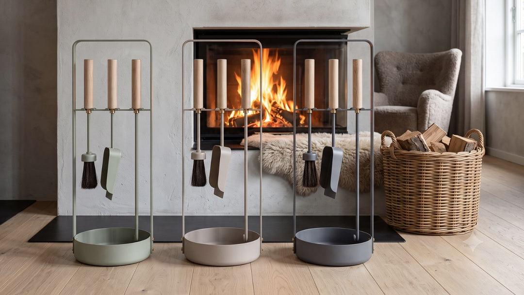 Three modern fireplace tool sets in sage, beige, and gray stand before a lit fireplace, with a wicker basket of logs and armchair nearby.
