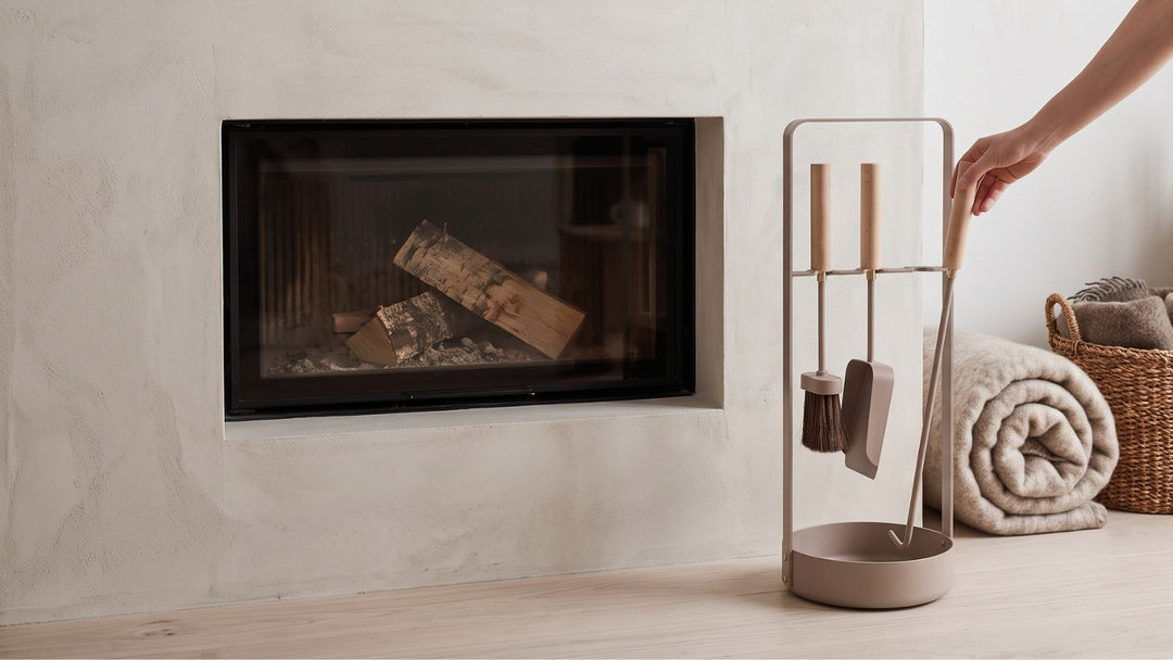 Modern fireplace with logs, minimalist metal and wood tool set, rolled blanket, and basket beside it.