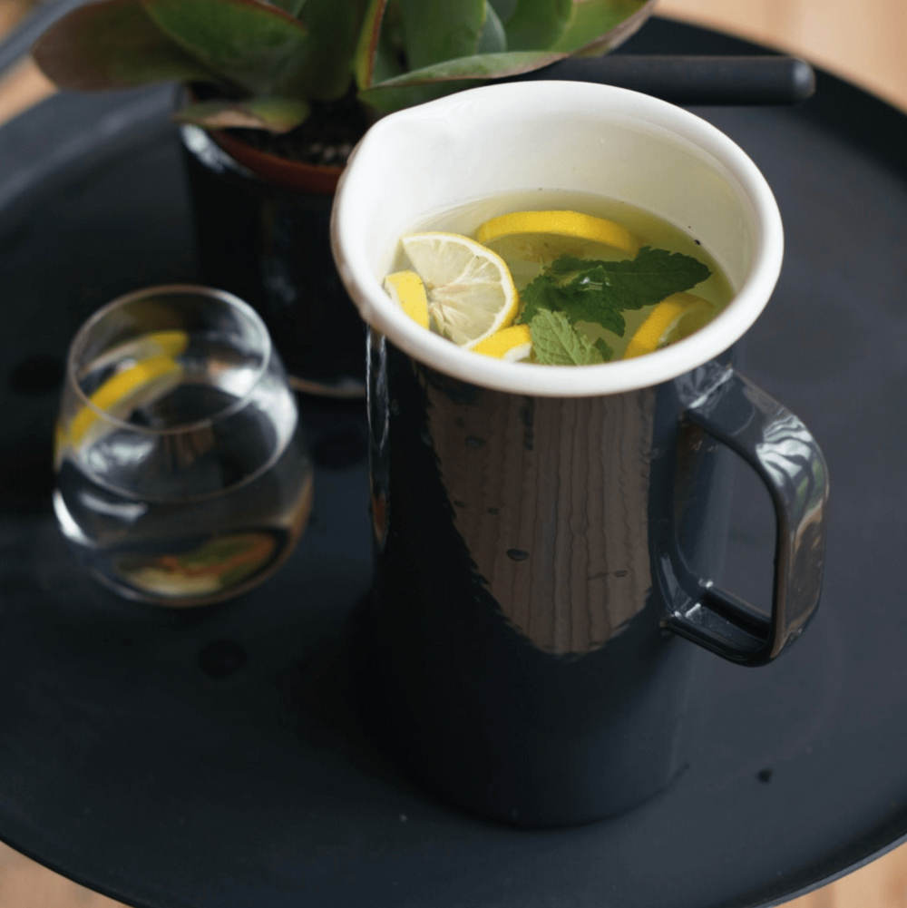 Emaille Krug 2L von LIVLIG, in Grau gehalten, mit Zitrone und Minze gefüllt, neben einem Glas auf einem schwarzen Tablett.
