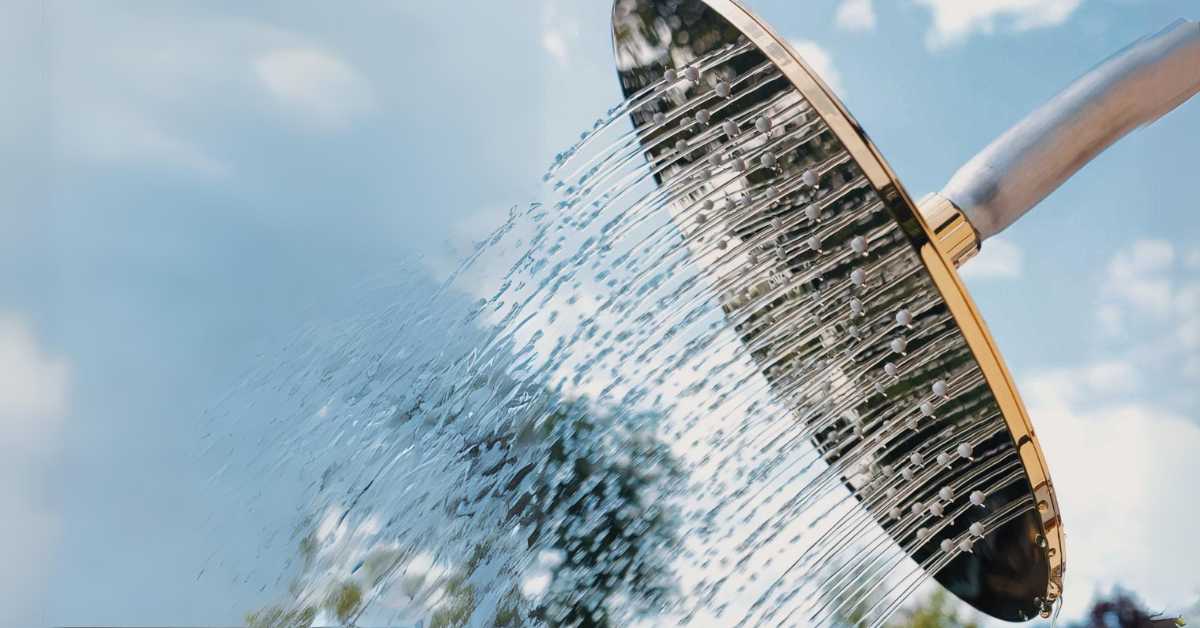 Eine Dusche mit goldenem Rand und silberfarbenem Arm sprüht Wasser vor einem blauen Himmel mit Wolken und Bäumen.