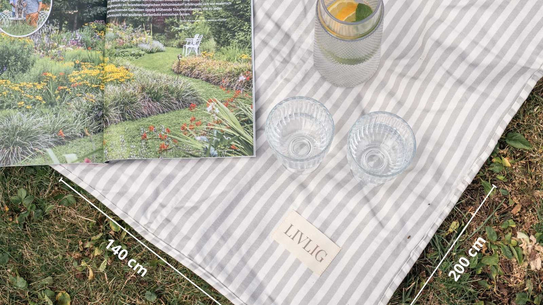Ein gestreiftes Picknicktuch liegt auf Gras. Darauf stehen eine Zeitschrift mit Gartenbildern, zwei Gläser und ein Krug mit Wasser. Text: "LIVLIG", "140 cm", "200 cm".