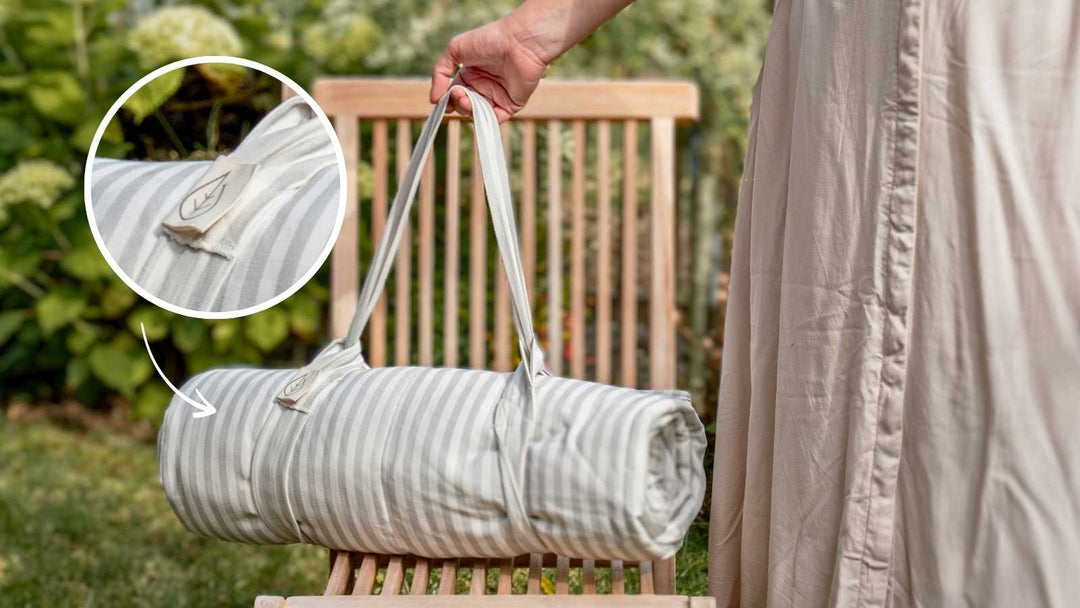 Eine aufgerollte, gestreifte Yogamatte mit Tragetasche wird von einer Person vor einem Garten gehalten. Ein eingenähter Anhänger mit Blattmotiv ist sichtbar.