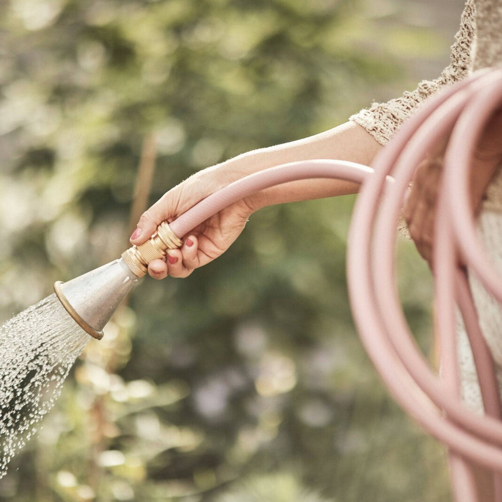 Rosafarbener Design-Gartenschlauch von LIVLIG, mit Brause in der Hand, Wassertropfen und grüner Hintergrund.