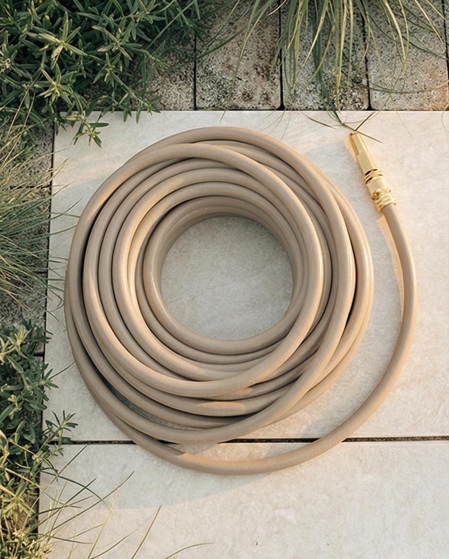 Draufsicht auf einen beigen Gartenschlauch mit goldenem Anschluss, der spiralförmig auf hellen Fliesen neben Grünpflanzen liegt.