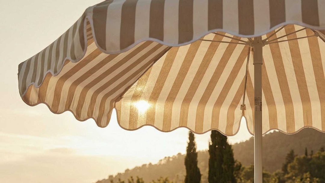 Ein gestreifter Sonnenschirm mit gewelltem Rand im Vordergrund, dahinter eine Landschaft mit sanftem Sonnenlicht. Beige und weiße Streifen dominieren.