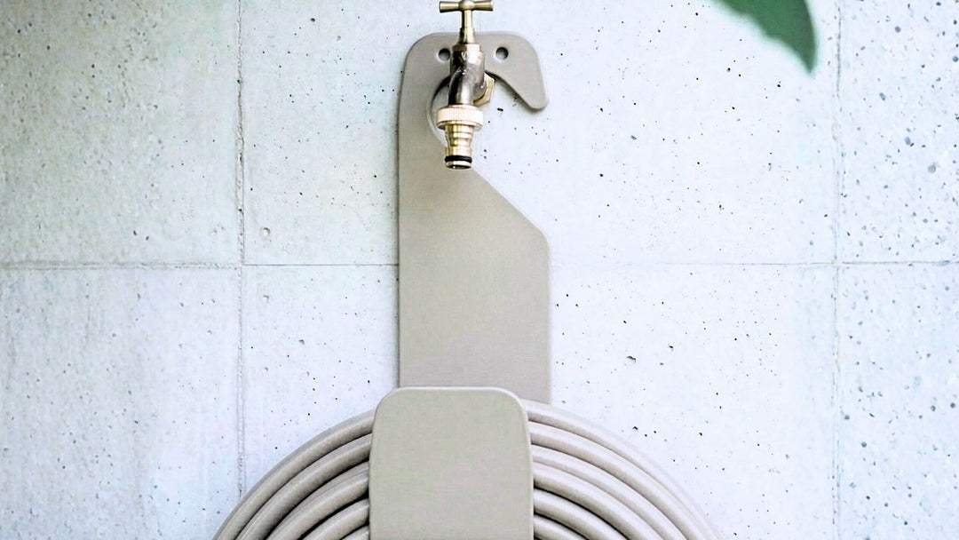 An einer hellen Betonwand mit Fugen hängt eine moderne, hellgraue Schlauchhalterung mit einem messingfarbenen Wasserhahn und einem darunter aufgerollten Gartenschlauch.