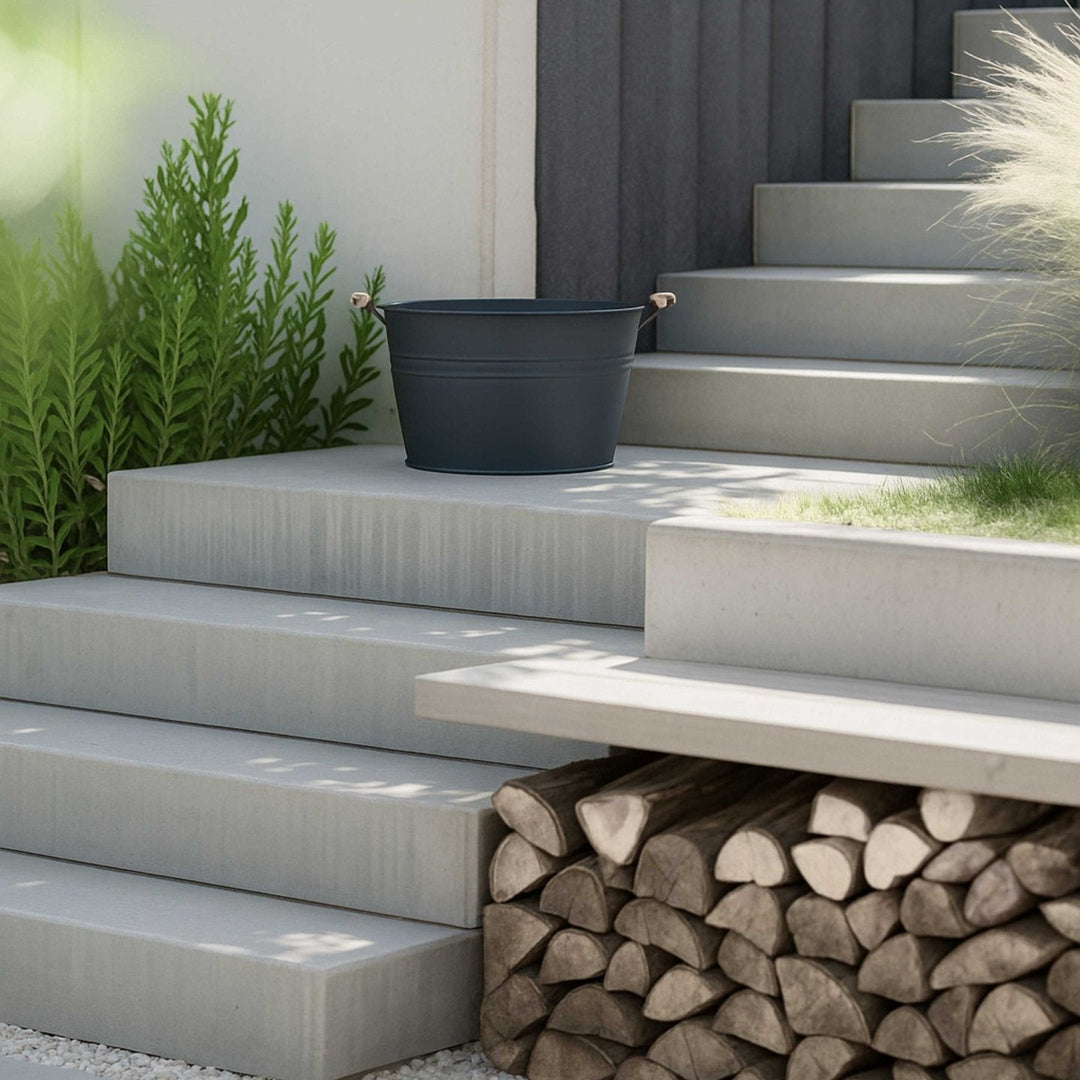Dunkelgraue Zinkwanne 30L von LIVLIG, auf Betonstufen neben grünen Pflanzen und einem Holzstapel platziert.