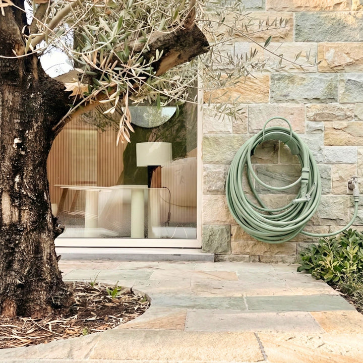 Grüner Design Gartenschlauch von LIVLIG, aufgerollt an einer Steinwand neben einem Baum.