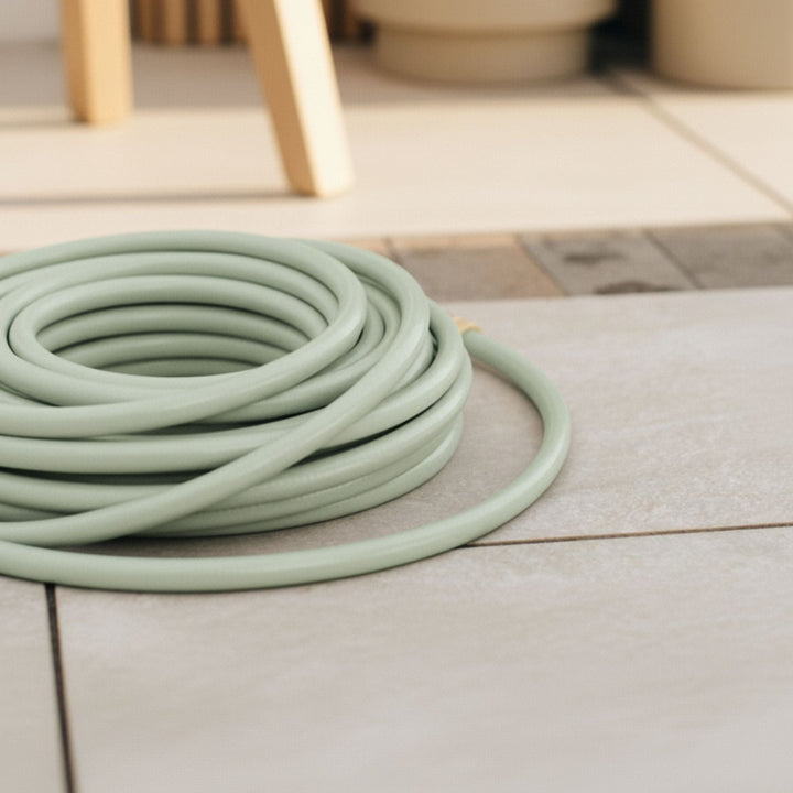Grüner Design Gartenschlauch Homefarming von LIVLIG, zusammengerollt auf hellen Steinplatten, mit einem Teil eines hellen Holzstuhls im Hintergrund.