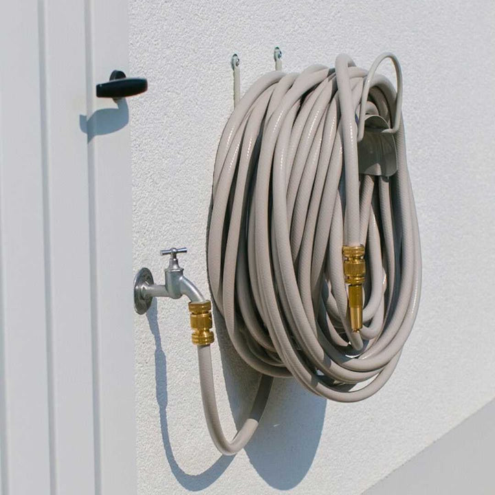 Ein aufgerollter, grauer Gartenschlauch hängt an einer weißen Wand neben einem Wasserhahn, der an einer weißen Tür befestigt ist.