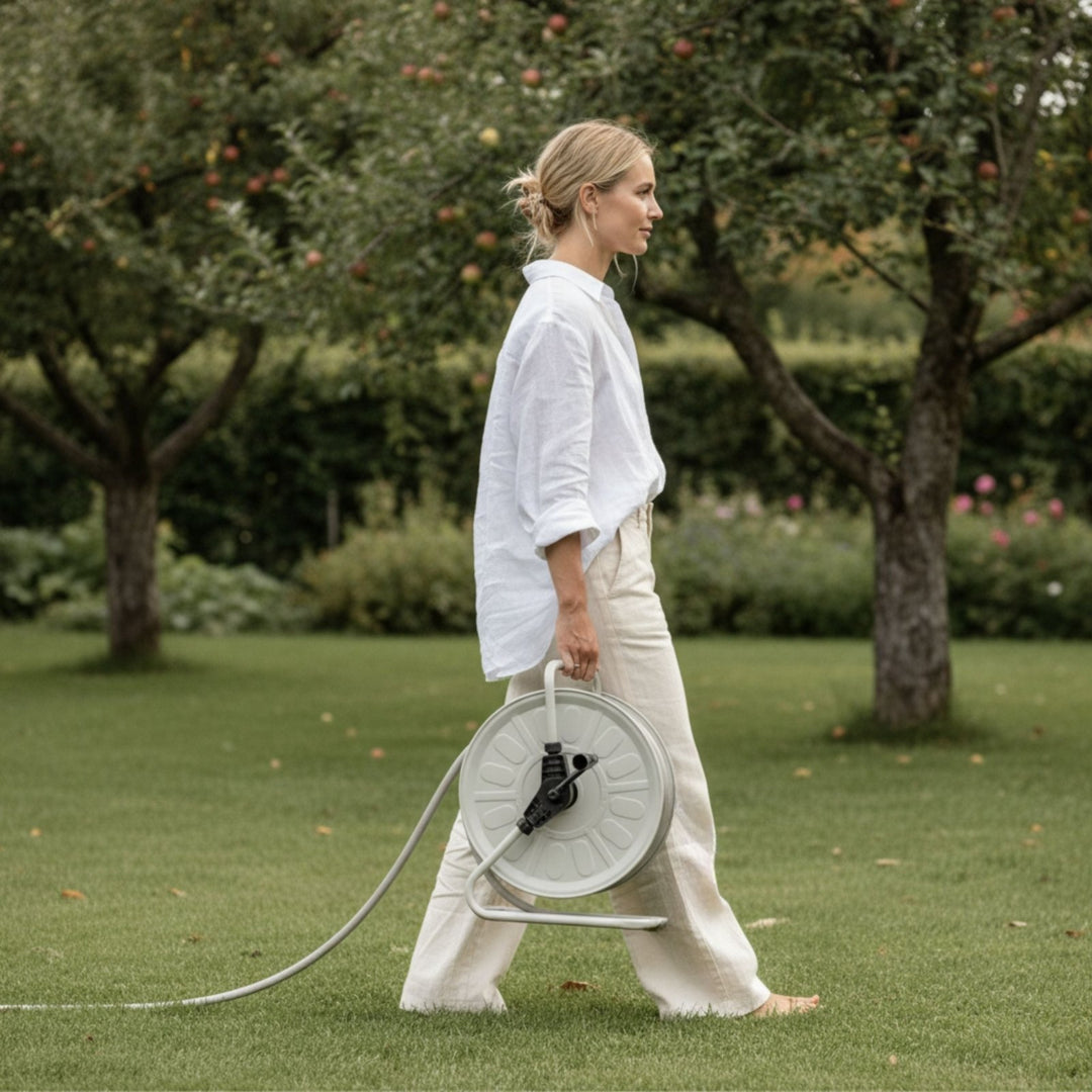 Gartenschlauch-Set mit Trommel in Greige von LIVLIG, von einer Frau barfuß über grüne Wiese getragen.