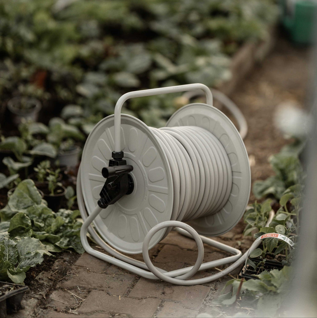 Eine graue Schlauchtrommel steht im Vordergrund, umgeben von Pflanzen auf einem Ziegelboden in einem Gewächshaus. Im Hintergrund weitere Pflanzen und unscharfe Töpfe.