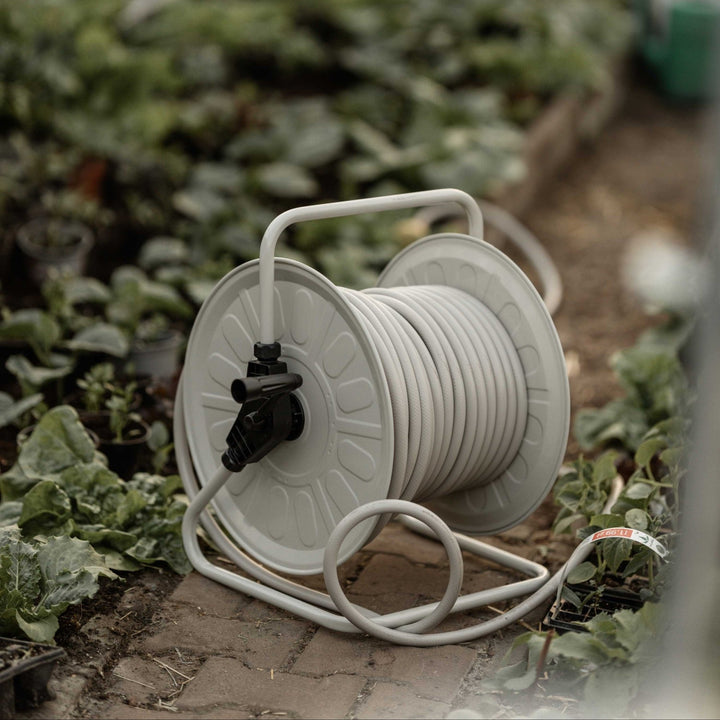 Eine graue Schlauchtrommel steht im Vordergrund, umgeben von Pflanzen auf einem Ziegelboden in einem Gewächshaus. Im Hintergrund weitere Pflanzen und unscharfe Töpfe.