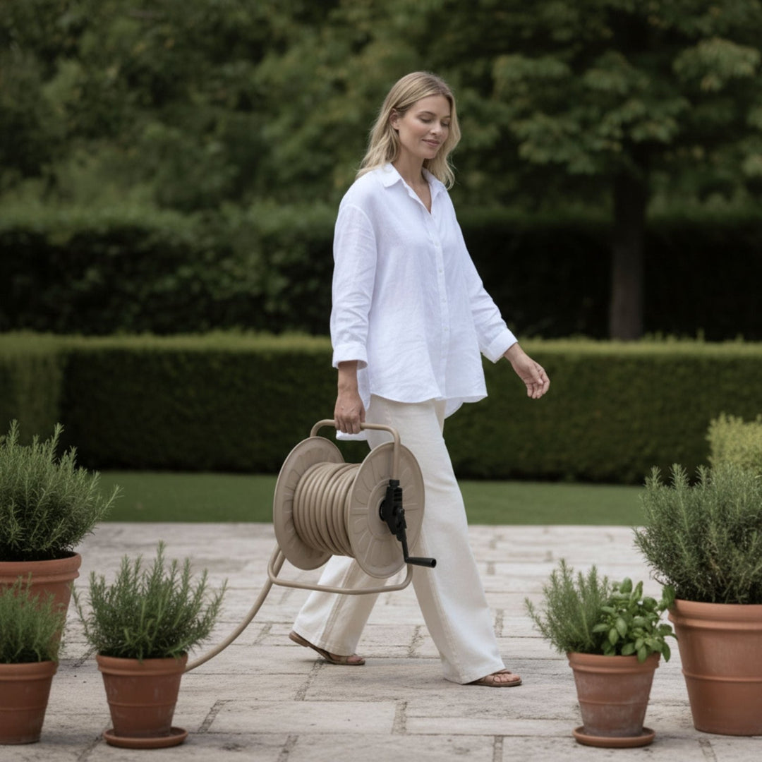 Beigefarbenes Gartenschlauch-Set mit Trommel von LIVLIG, von einer Frau in weißem Hemd über eine Terrasse getragen.