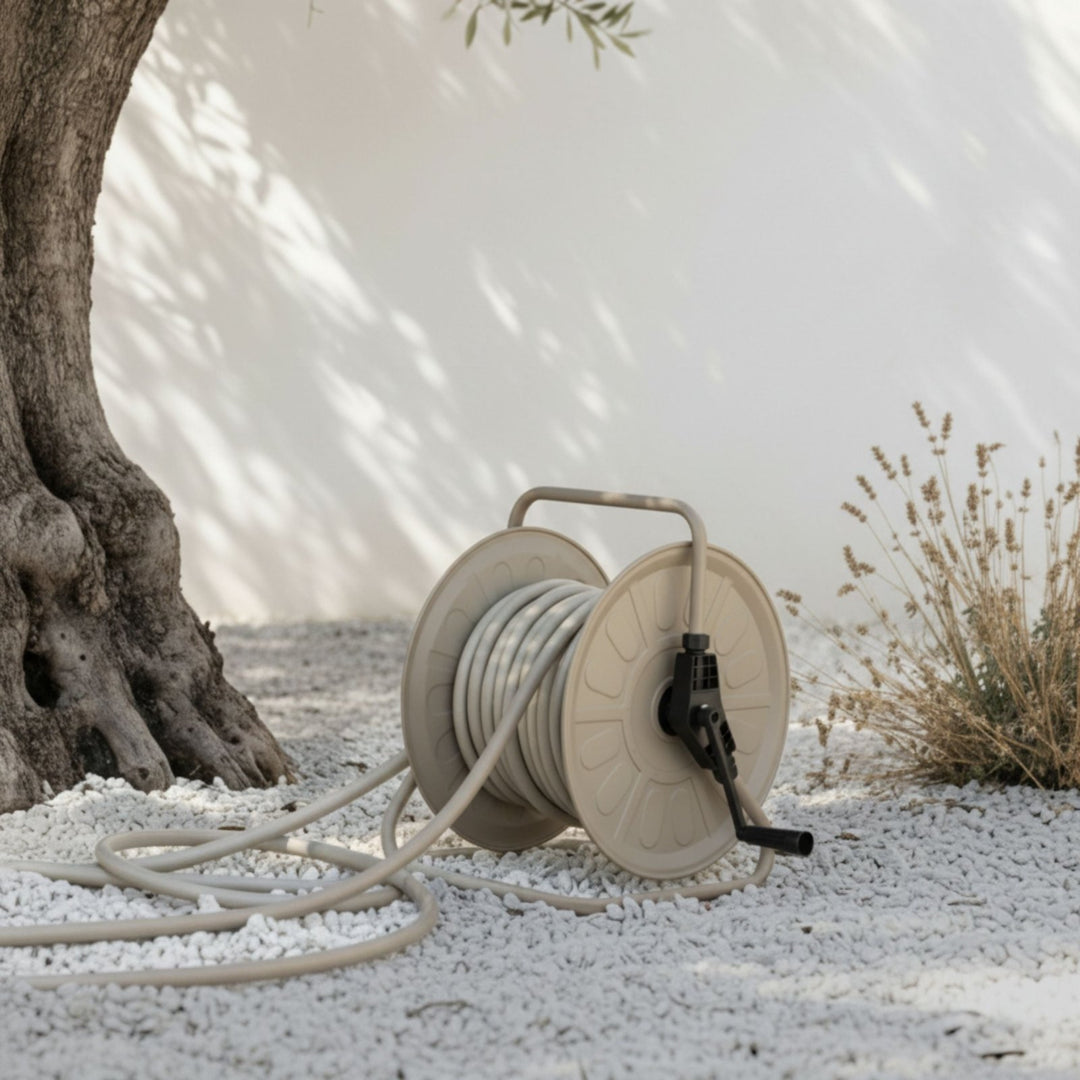Gartenschlauch Set mit Trommel in Sandy Stone von LIVLIG auf weißem Kies, neben einem Baumstamm und trockenen Pflanzen, mit Schatten auf einer weißen Wand.