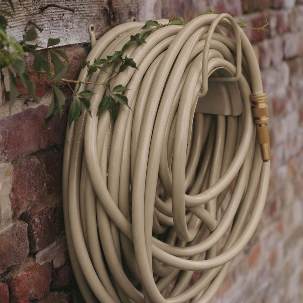 Gartenschlauch Set in Sandy Stone von LIVLIG, sauber aufgerollt mit Messingdüse, vor einer Ziegelwand und Efeu.