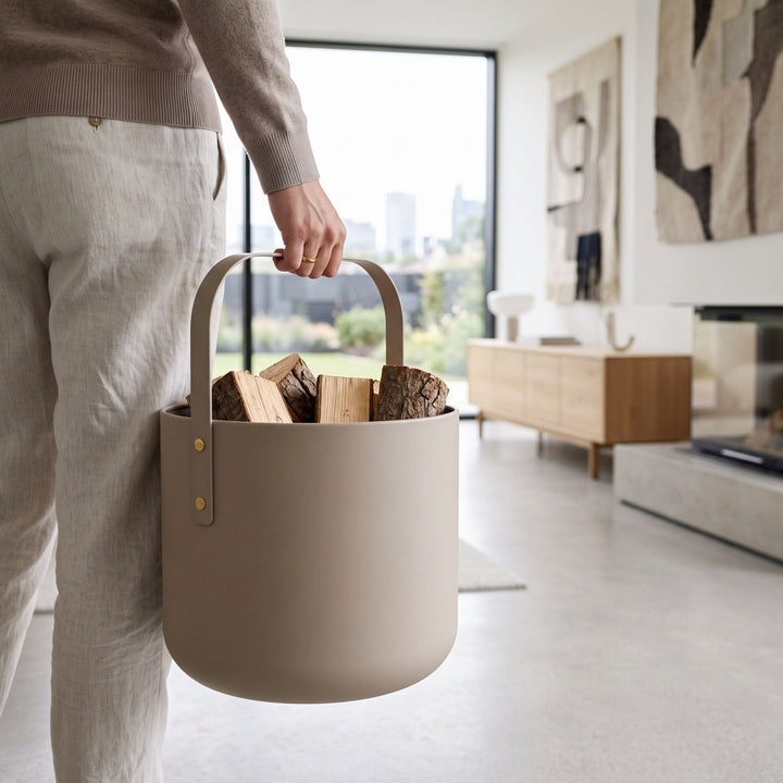 Eleganter metallener Kaminholzkorb in taupe von LIVLIG, mit geschwungener Henkel und goldfarbenen Beschlägen, gefüllt mit Holzscheiten, im Wohnzimmer gezeigt.