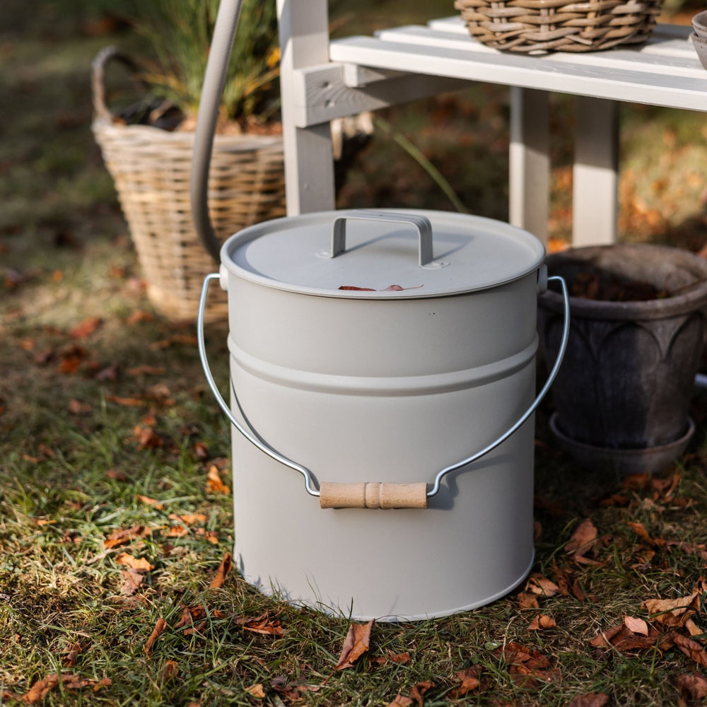 Grauer Metalleimer I Ascheeimer mit Deckel von LIVLIG, mit Holzgriff und im Freien auf Gras mit Herbstlaub stehend.
