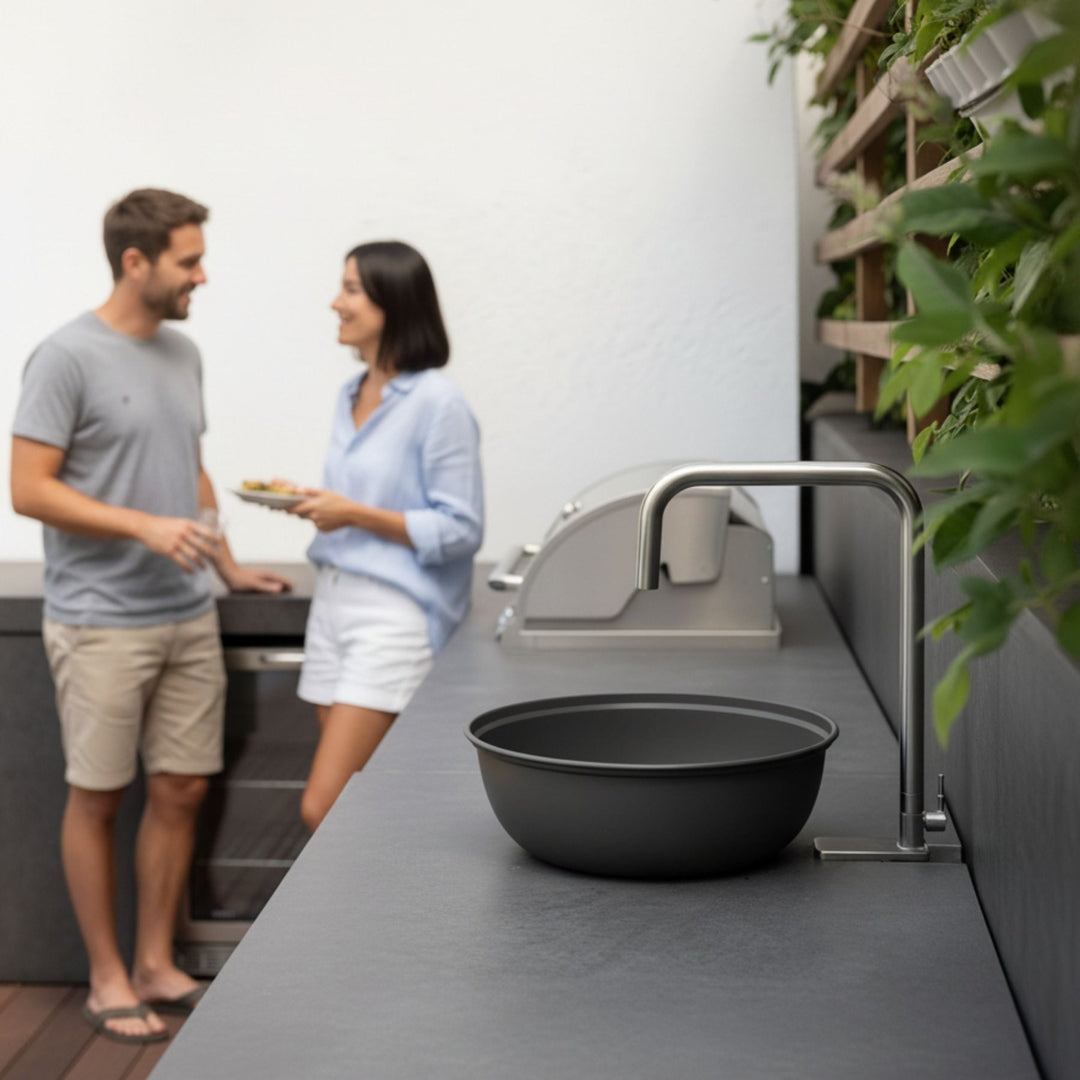 Schwarzer Wasserhahn von LIVLIG auf einer grauen Arbeitsplatte im Außenbereich mit Pflanzwand und zwei Personen im Hintergrund.