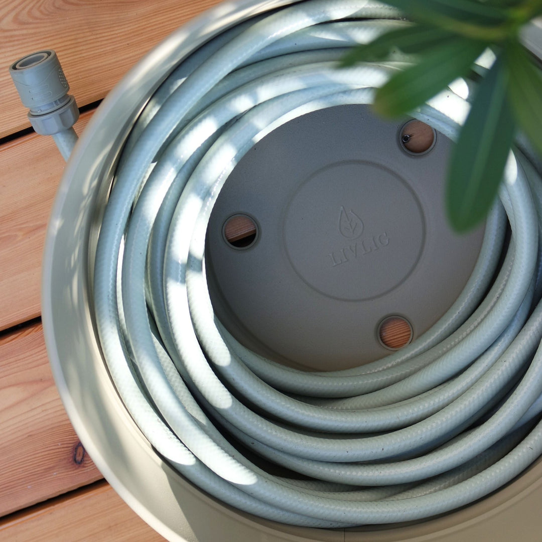 Metall-Schlauchtopf von LIVLIG mit aufgerolltem Gartenschlauch und Anschlussstück in einem runden, beigen Topf.