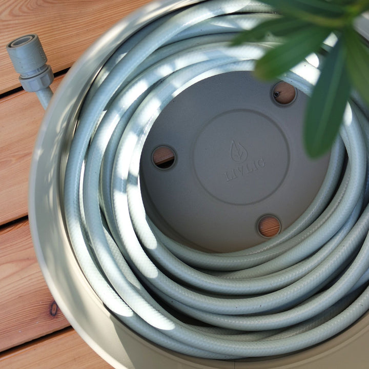 Metall-Schlauchtopf von LIVLIG mit aufgerolltem Gartenschlauch und Anschlussstück in einem runden, beigen Topf.