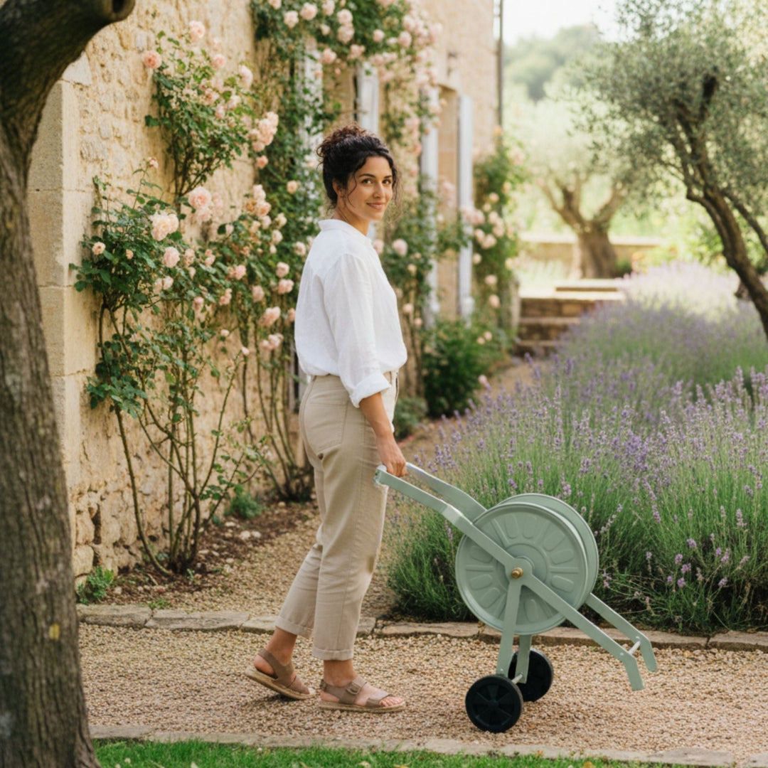 Grüner Schlauchwagen Komplettset Homefarming von LIVLIG, mit schwarzen Rädern, vor einer Frau und blühenden Lavendel im Garten.
