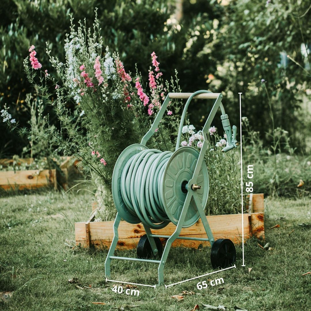 Grüner Schlauchwagen Komplettset Homefarming von LIVLIG, vor einem Beet mit rosafarbenen und weißen Blumen.