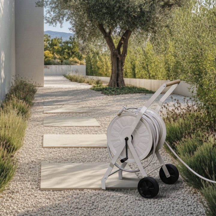 Weißer Metall-Schlauchwagen von LIVLIG mit zwei Rädern und Holzgriff auf Kiesweg mit Trittplatten.