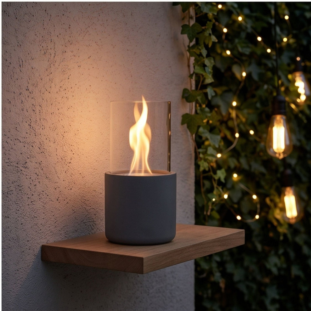 Gray cylindrical outdoor tabletop fireplace with clear glass enclosure and flickering flame, placed on wooden shelf beside ivy wall with string lights.