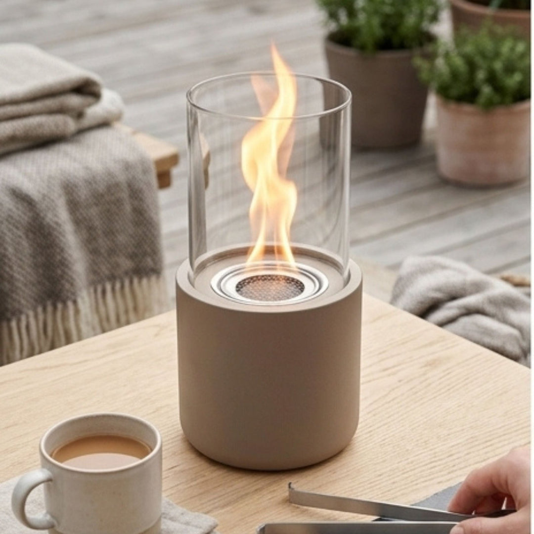Beige cylindrical outdoor tabletop fireplace with clear glass wind guard and visible flame, placed on wooden table with ceramic mug and folded blanket nearby.