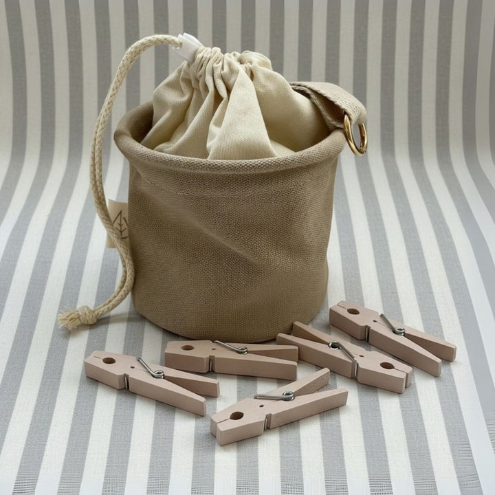 Wäscheklammerbeutel mit 50 Wäscheklammern von LIVLIG, aus beigem Stoff, mit Kordelzug und goldenem Ring, umgeben von Holzwäscheklammern auf gestreiftem Untergrund.
