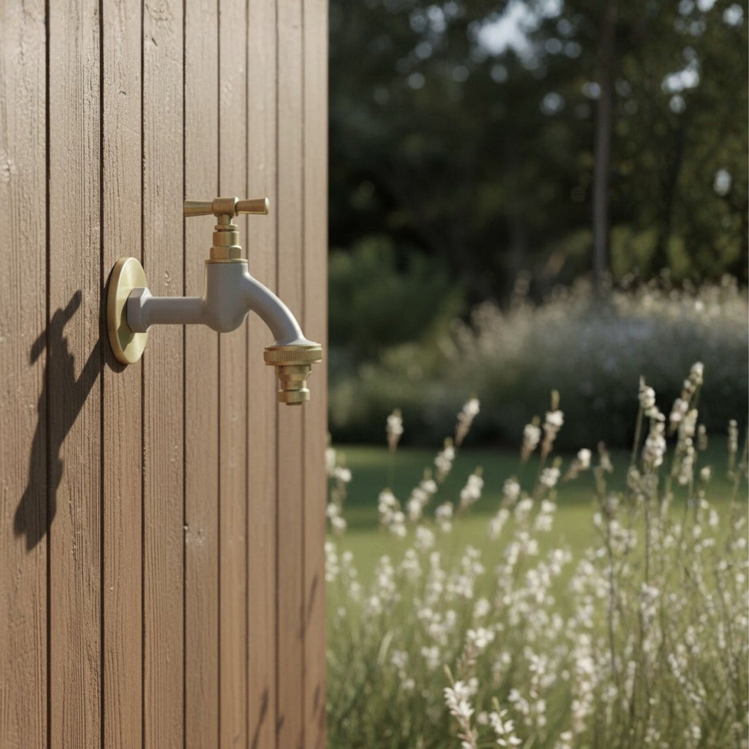 Grauer Gartenwasserhahn mit goldenen Akzenten und Rosettengarnitur von LIVLIG, montiert an brauner Holzverkleidung mit unscharfem Gartenhintergrund.
