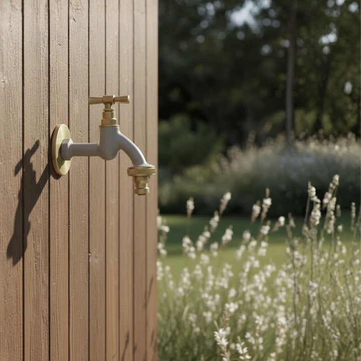 Grauer Gartenwasserhahn mit goldenen Akzenten und Rosettengarnitur von LIVLIG, montiert an brauner Holzverkleidung mit unscharfem Gartenhintergrund.